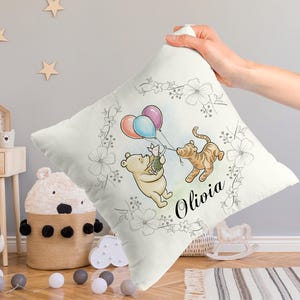 May include: A white throw pillow with a watercolor illustration of Winnie the Pooh, Piglet, and Tigger holding balloons. The illustration is surrounded by a floral wreath. The name Olivia is written in black script below the illustration.