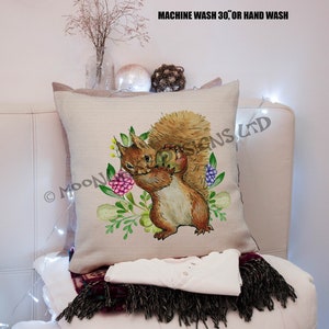 May include: A beige cushion featuring a watercolour illustration of a squirrel holding a nut. The squirrel is surrounded by green leaves and pink flowers. The text "MACHINE WASH 30°, OR HAND WASH" is printed in black at the top of the cushion.