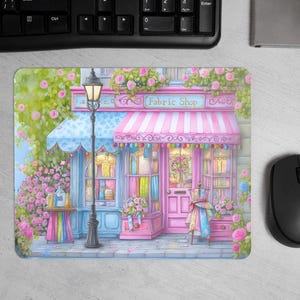May include: A mousepad featuring a watercolour illustration of a fabric shop. The shop has a pink and blue facade, a striped awning, and the text "Fabric Shop". The scene includes flowers and a vintage street lamp.