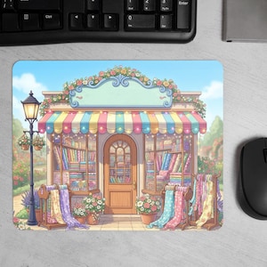May include: A mouse mat with a whimsical illustration of a shop with a striped awning and floral decorations. The shop displays fabrics and is set against a blue sky. A black computer mouse and keyboard are in the background.