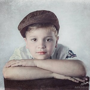 May include: A young boy with short blond hair wearing a brown cap and a white shirt with a blue patch on the sleeve, rests his chin on his folded arms, looking thoughtfully at the camera.
