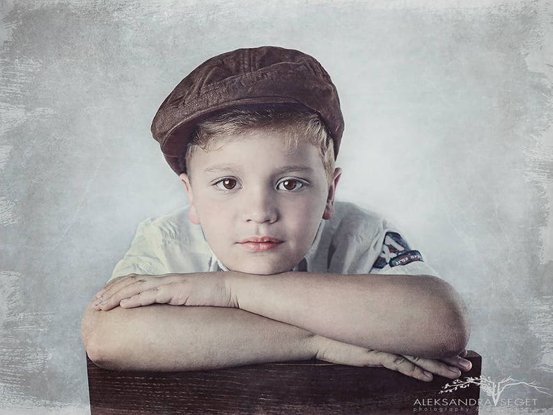 May include: A young boy with short blond hair wearing a brown cap and a white shirt with a blue patch on the sleeve, rests his chin on his folded arms, looking thoughtfully at the camera.