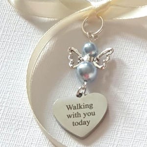 Puede incluir: Un dije de ángel plateado con cuentas de perlas azules y delicadas alas, suspendido de una cinta color crema. Una etiqueta en forma de corazón debajo dice "Walking with you today". Un recuerdo sentimental.