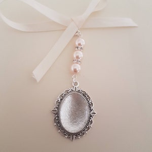 May include: A silver-toned oval pendant with an ornate filigree border, suspended from a white ribbon. The pendant is attached to a vertical strand featuring three pale pink faux pearls separated by two small silver-toned rings with clear crystals.