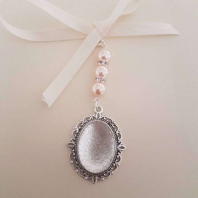 May include: A silver-toned oval pendant with an ornate filigree border, suspended from a white ribbon. The pendant is attached to a vertical strand featuring three pale pink faux pearls separated by two small silver-toned rings with clear crystals.