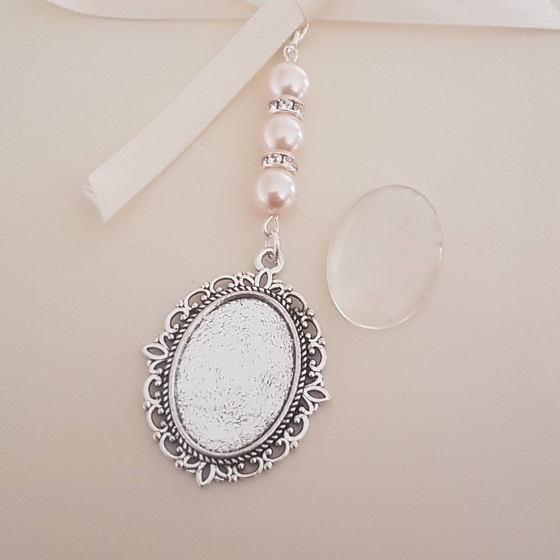 May include: An ornate silver-toned oval pendant with a decorative frame, suspended from a strand of pink pearls and clear rhinestones. A cream-colored ribbon and a clear oval glass piece are also present. The pendant is likely for a necklace or charm.