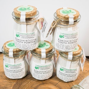 Peut inclure: Quatre bocaux en verre avec des couvercles en liège remplis de poudre blanche étiquetés "Remineralizing Tooth Powder" avec un logo de feuille verte. Les bocaux sont disposés en cercle sur une surface en bois avec une brosse à dents en bambou, de l'anis étoilé, des clous de girofle et une branche de menthe.