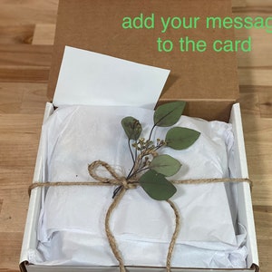May include: A white gift box with a brown lid is open, revealing a white tissue paper-wrapped gift. The tissue paper is tied with twine and decorated with a sprig of green leaves. The lid of the box has the text "add your message to the card" written on it.
