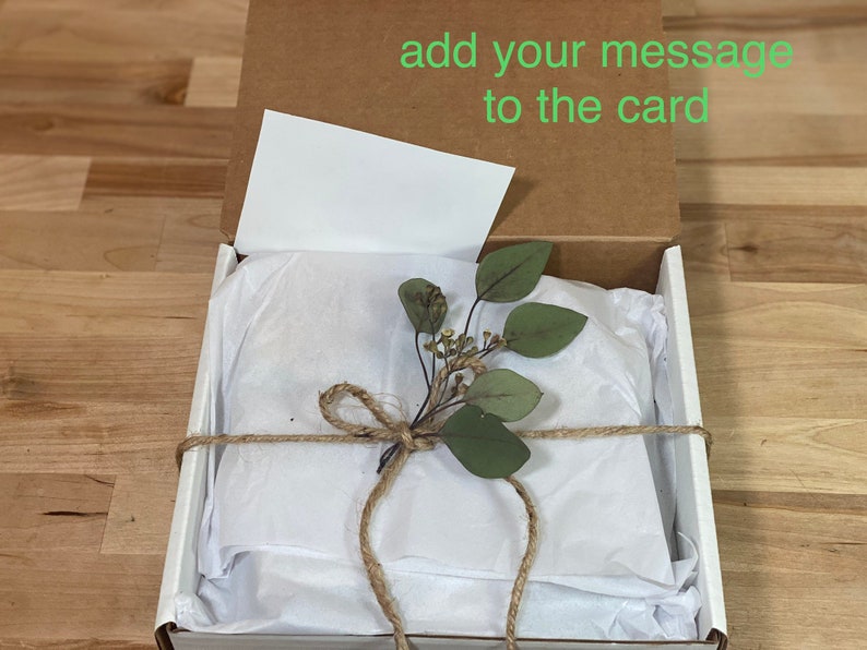 May include: A white gift box with a brown lid is open, revealing a white tissue paper-wrapped gift. The tissue paper is tied with twine and decorated with a sprig of green leaves. The lid of the box has the text "add your message to the card" written on it.