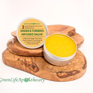 Peut inclure: Un petit pot de baume infusé au gingembre et au curcuma de couleur jaune sur une surface en bois. Le pot est muni d'une étiquette blanche avec le texte "GreenLife Apothecary", "greenlifeapothecary.com", "craftimamma", "Gingembre & Curcuma Infusé en Baume", et "Elaboré avec du gingembre et du curcuma, lentement infusés dans des huiles nourrissantes pour capturer la pleine vitalité de ces plantes".