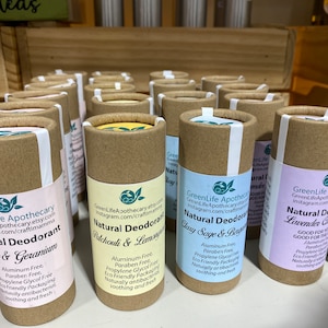 Op de afbeelding: Een display met natuurlijke deodorant tubes in verschillende geuren, waaronder Rozen & Geranium, Patchouli & Citroengras, Salie & Bergamot en Lavendel & Kamille. De tubes zijn gemaakt van karton en hebben een bruin-witte kleurstelling. De labels tonen de merknaam "GreenLife Apothecary" en vermelden de ingredi&euml;nten en voordelen van elke geur.