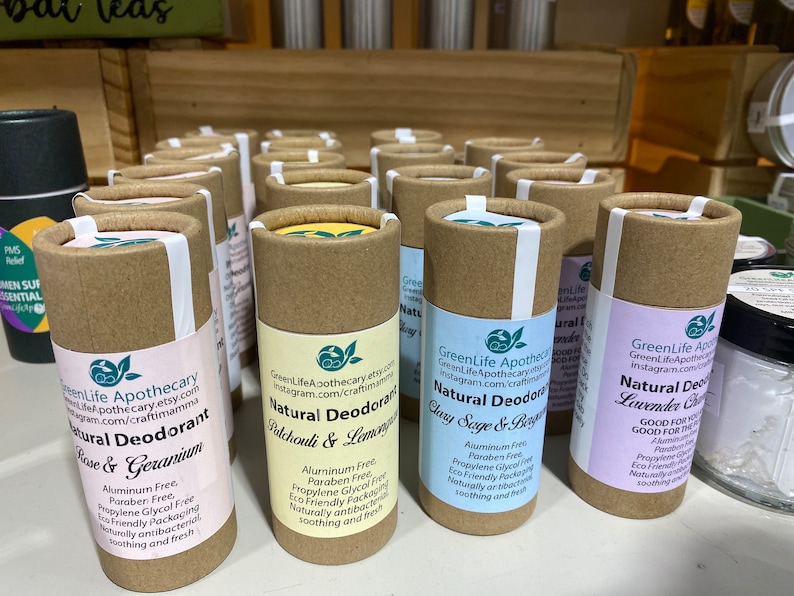 Op de afbeelding: Een display met natuurlijke deodorant tubes in verschillende geuren, waaronder Rozen & Geranium, Patchouli & Citroengras, Salie & Bergamot en Lavendel & Kamille. De tubes zijn gemaakt van karton en hebben een bruin-witte kleurstelling. De labels tonen de merknaam "GreenLife Apothecary" en vermelden de ingredi&euml;nten en voordelen van elke geur.