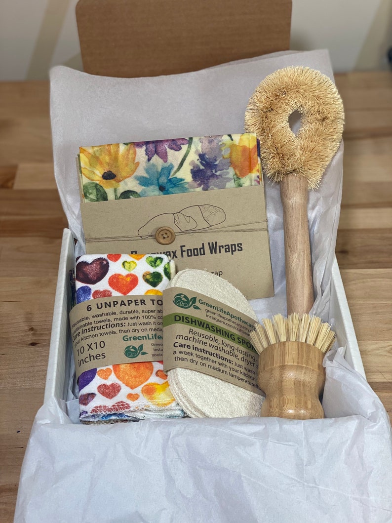 May include: A gift box filled with eco-friendly kitchen supplies. The box contains six unpaper towels, two reusable food wraps, a natural sponge, and a wooden dish brush. The unpaper towels are printed with colorful hearts and the food wraps have a floral design. The text on the packaging reads "6 Unpaper Towels" and "Wax Food Wraps".