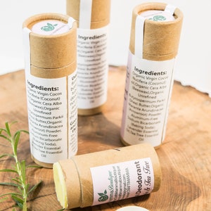 Op de afbeelding: Drie bruine kartonnen buizen met witte etiketten die natuurlijke deodorant bevatten. De etiketten vermelden de ingredi&euml;nten, waaronder biologische kokosolie, sheaboter en tea tree etherische olie. De buizen staan op een houten oppervlak met een takje rozemarijn.