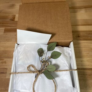 May include: A white gift box with a brown twine bow and a sprig of green eucalyptus leaves. The box is filled with white tissue paper.