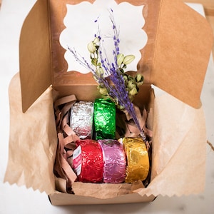 Puede incluir: Una caja de cartón marrón con una ventana que muestra una rama de flores moradas. La caja contiene cinco bombas de baño envueltas en papel de aluminio plateado, verde, rojo, rosa y dorado.