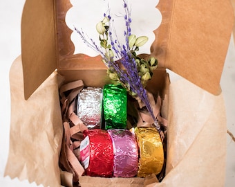 May include: A brown cardboard box with a window showing a sprig of purple flowers. The box contains five wrapped bath bombs in silver, green, red, pink, and gold foil.