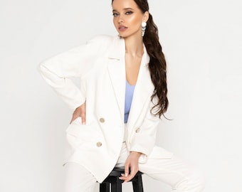 May include: A white blazer with a double-breasted closure and a white pair of pants. The blazer has gold buttons and the model is wearing a light blue top underneath.