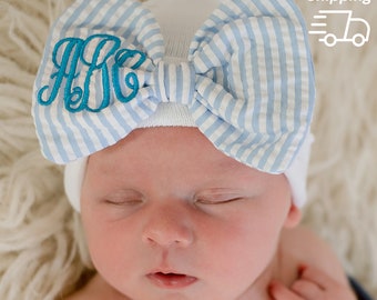 May include: A white baby hat with a blue and white striped bow. The bow has a turquoise monogram "ABC" embroidered on it.