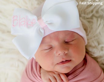 May include: A newborn baby wearing a pink beanie with a large white bow. The bow has the name "Blake" embroidered in pink. The baby is wrapped in a pink blanket and is lying on a white, fluffy surface.