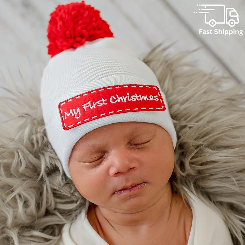 May include: A white knit baby beanie with a red pom-pom on top. The beanie has a red rectangular patch with the embroidered text "My First Christmas". The baby is lying on a soft, gray surface.
