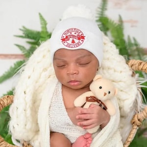 May include: A newborn baby wearing a white knit hat with a red "North Pole Special Delivery" patch, sleeping with a cream-colored teddy bear.