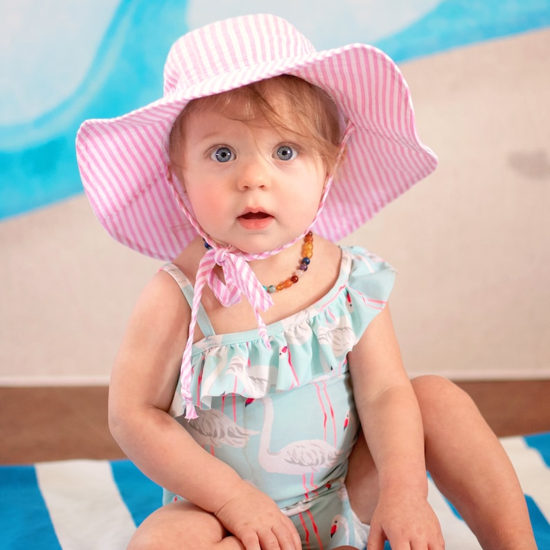May include: A baby wearing a pink and white striped sun hat with a matching tie. The baby is also wearing a light blue swimsuit with a flamingo pattern and a colorful beaded necklace.