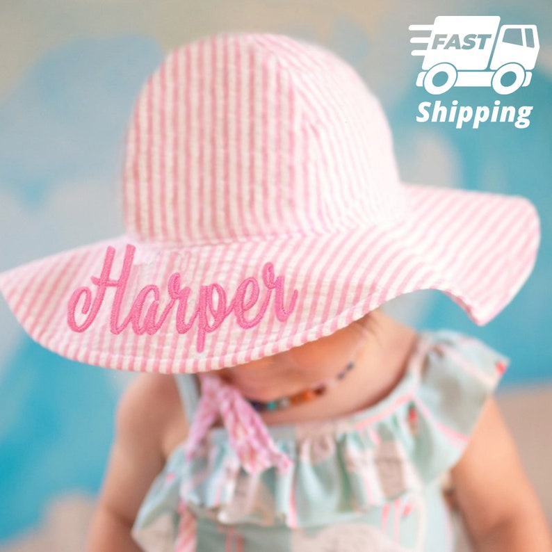 May include: A pink and white striped sun hat with the name "Harper" embroidered in pink script across the brim. The hat has a wide brim and is designed for a child.