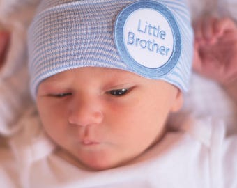May include: A newborn baby wearing a light blue and white striped beanie with a circular patch. The patch has the words "Little Brother" embroidered in blue. The baby is wearing a white onesie.