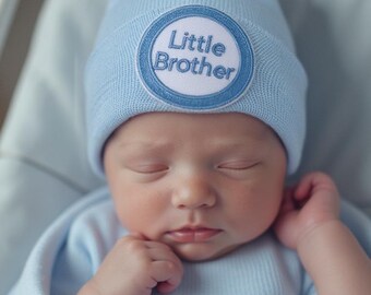 May include: A newborn baby wearing a light blue beanie with a circular patch that reads "Little Brother" in dark blue lettering. The baby is also wearing a light blue onesie and is sleeping peacefully.