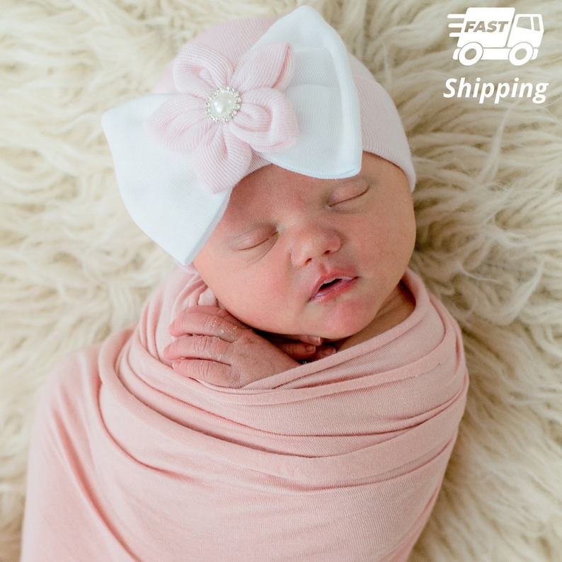 May include: A pink and white baby hat with a large pink flower and a pearl-like embellishment. The hat is on a baby's head, who is sleeping.