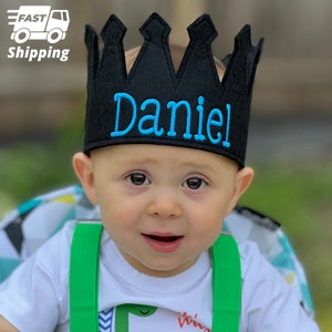 May include: A black felt crown with blue embroidery that says "Daniel". The crown is on a baby's head.