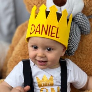 Puede incluir: Un beb&eacute; sonriente con una corona amarilla con el nombre "DANIEL" en letras rojas. El beb&eacute; tambi&eacute;n lleva un body blanco con un gr&aacute;fico de corona dorada y las palabras "Mr. ONE". Tirantes negros visibles.