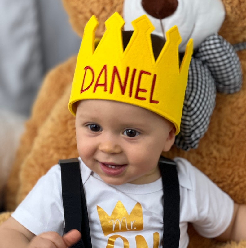Puede incluir: Un beb&eacute; sonriente con una corona amarilla con el nombre "DANIEL" en letras rojas. El beb&eacute; tambi&eacute;n lleva un body blanco con un gr&aacute;fico de corona dorada y las palabras "Mr. ONE". Tirantes negros visibles.