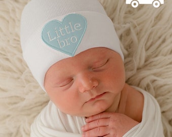 May include: A white baby hat with a blue heart that says "Little bro" on it. The hat is on a baby's head.