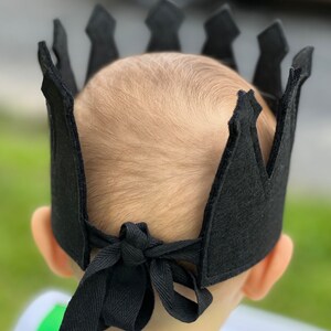 May include: A black felt crown with a pointed top and a tie at the back. The crown is worn on a child's head.