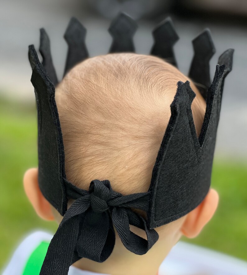 May include: A black felt crown with a pointed top and a tie at the back. The crown is worn on a child's head.