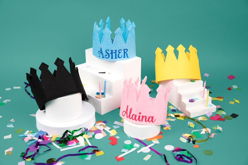 May include: Four felt crowns with names embroidered on them. The crowns are blue, black, pink, and yellow. The crowns are on white platforms with confetti and streamers scattered around them.