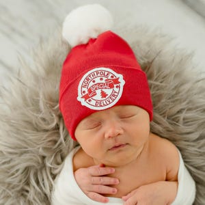 May include: A red knit baby hat with a white pom pom and a white embroidered patch that says "North Pole Special Delivery".