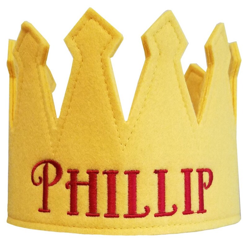 Puede incluir: Una corona de fieltro amarillo con el nombre "PHILLIP" bordado en rojo. La corona tiene arcos puntiagudos y un dise&ntilde;o cosido. Adecuada para una fiesta de cumplea&ntilde;os o disfraz.