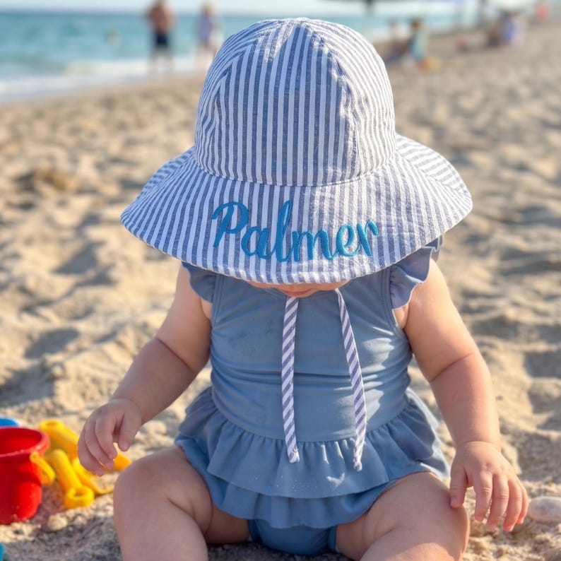 May include: A baby wearing a blue swimsuit and a striped sun hat with the name "Palmer" embroidered in blue. The hat has a wide brim and a chin strap. The baby is sitting on a sandy beach.
