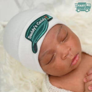 May include: White baby hat with a green and white embroidered patch that says "Daddy's Caddy" and a golf club icon.