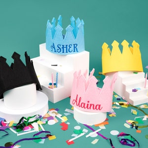 Puede incluir: Cuatro coronas de fieltro de colores, incluyendo azul con "Asher", rosa con "Alaina", amarillo y negro, sobre pedestales blancos. Confeti y velas a&ntilde;aden al tema festivo de cumplea&ntilde;os.