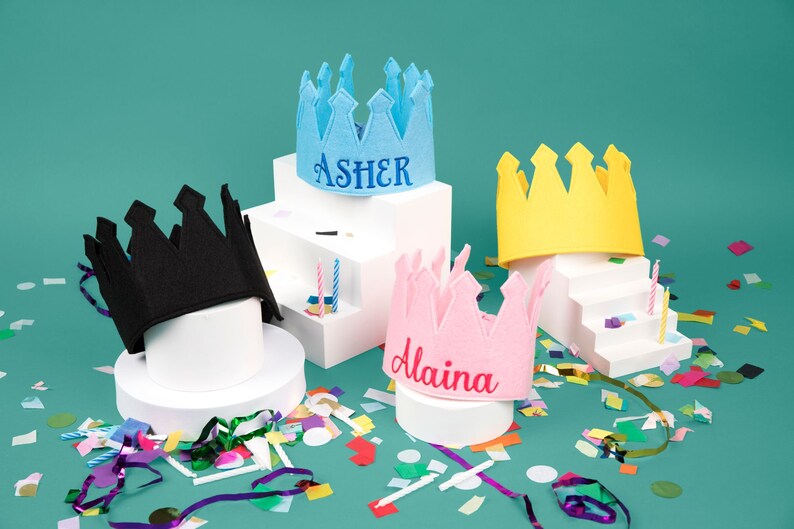 Puede incluir: Cuatro coronas de fieltro de colores, incluyendo azul con "Asher", rosa con "Alaina", amarillo y negro, sobre pedestales blancos. Confeti y velas a&ntilde;aden al tema festivo de cumplea&ntilde;os.