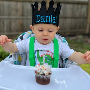 May include: A baby wearing a black crown with the name "Daniel" embroidered on it, sits in a high chair with a cupcake with a single lit candle in front of them.