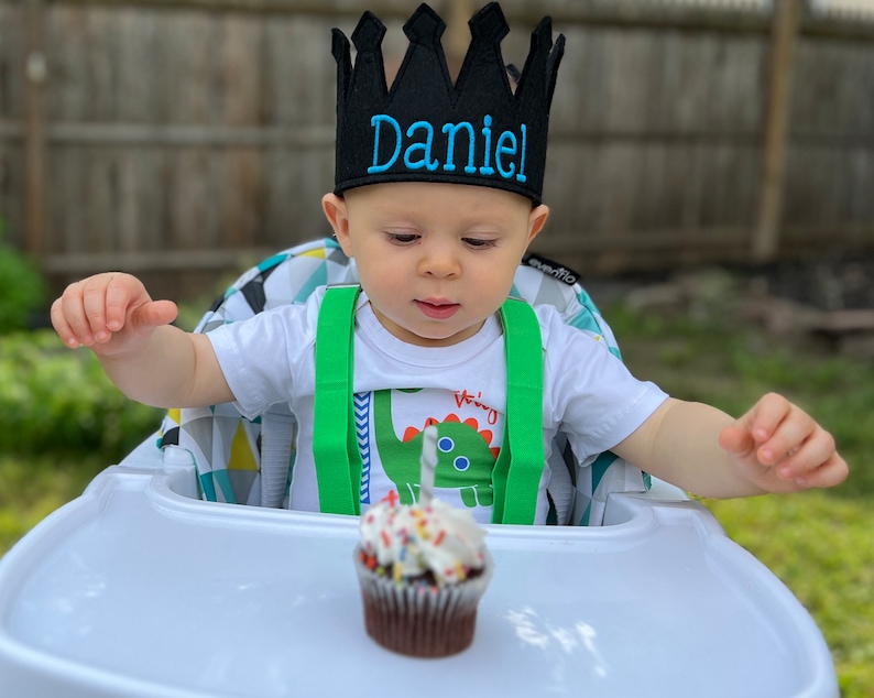 May include: A baby wearing a black crown with the name "Daniel" embroidered on it, sits in a high chair with a cupcake with a single lit candle in front of them.