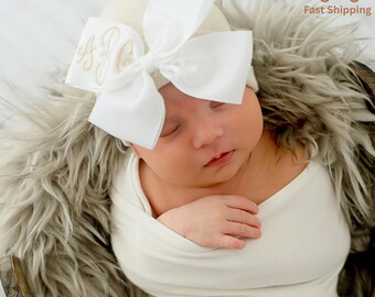 May include: A newborn baby wrapped in a white blanket, wearing a cream-colored beanie with a large white bow. The bow has embroidered initials. The baby is nestled in a wooden basket lined with faux fur.