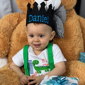 May include: A baby boy wearing a black crown with the name "Daniel" embroidered in blue thread. He is wearing a white shirt with a green dinosaur graphic and the text "It's my 1st" and green suspenders. He is sitting on a brown teddy bear.