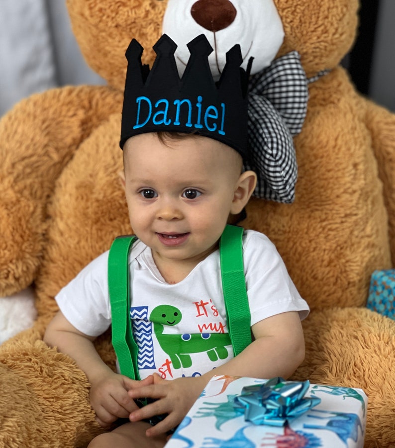 May include: A baby boy wearing a black crown with the name "Daniel" embroidered in blue thread. He is wearing a white shirt with a green dinosaur graphic and the text "It's my 1st" and green suspenders. He is sitting on a brown teddy bear.
