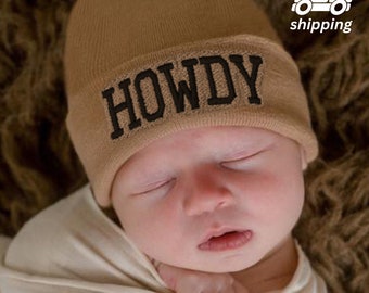 May include: A brown knitted baby hat with the word "HOWDY" embroidered in black.